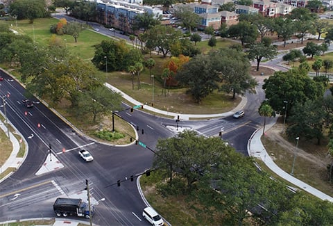 Cars drive past a tree-shaded raised median seen from above. 
