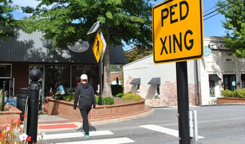 A pedestrian crosses in a crosswalk with a yellow pedestrian crossing sign in the foreground.