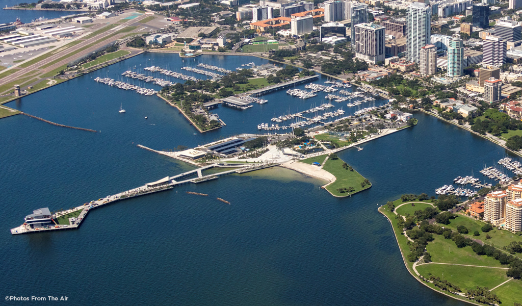 An aerial color photo of the city of St. Petersburg, Florida, shows tall buildings in an urban downtown area located close to Tampa Bay shoreline and the extensive tree canopy of South St. Petersburg peninsula.