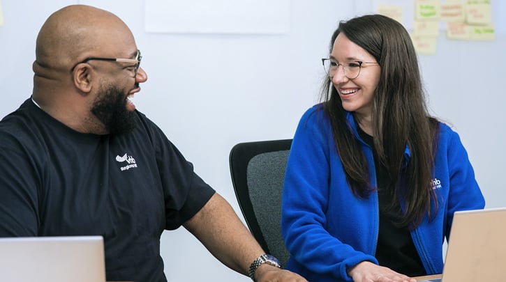 Two people sitting at a table with laptops, smiling at each other, with sticky notes on the wall behind them.