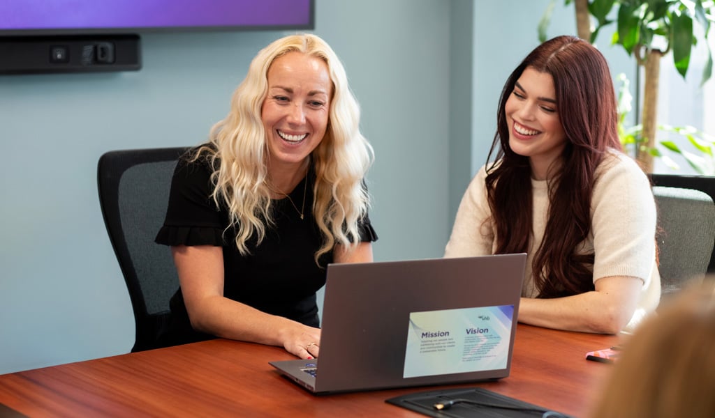 Two women smiling and looking at a laptop in a conference room.