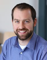 Andrew is seated at a conference table in a blue button-up shirt with chairs and a large window in the background.