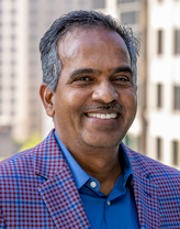 babuji smiling outside in a checkered blazer and blue shirt.