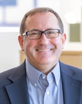 A man in a navy blazer and light blue dress shirt seated in a bright modern office with plants in the background.