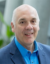 Joe wearing a navy blazer and blue shirt in front of a blurred background.