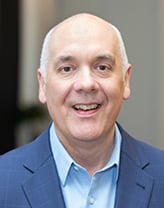 Joe wearing a navy blazer and blue shirt in front of a blurred background.