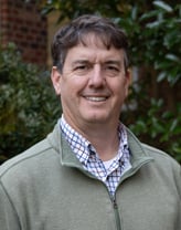 A man in a light green quarter-zip and plaid shirt standing outside in front of bushes and a brick building.