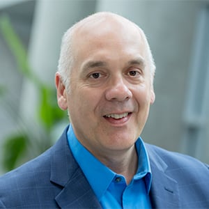 Joe wearing a navy blazer and blue shirt in front of a blurred background.