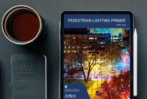 Next to a pen, coffee cup, and notebook, an iPad on a desk features the Pedestrian Lighting Primer screen.