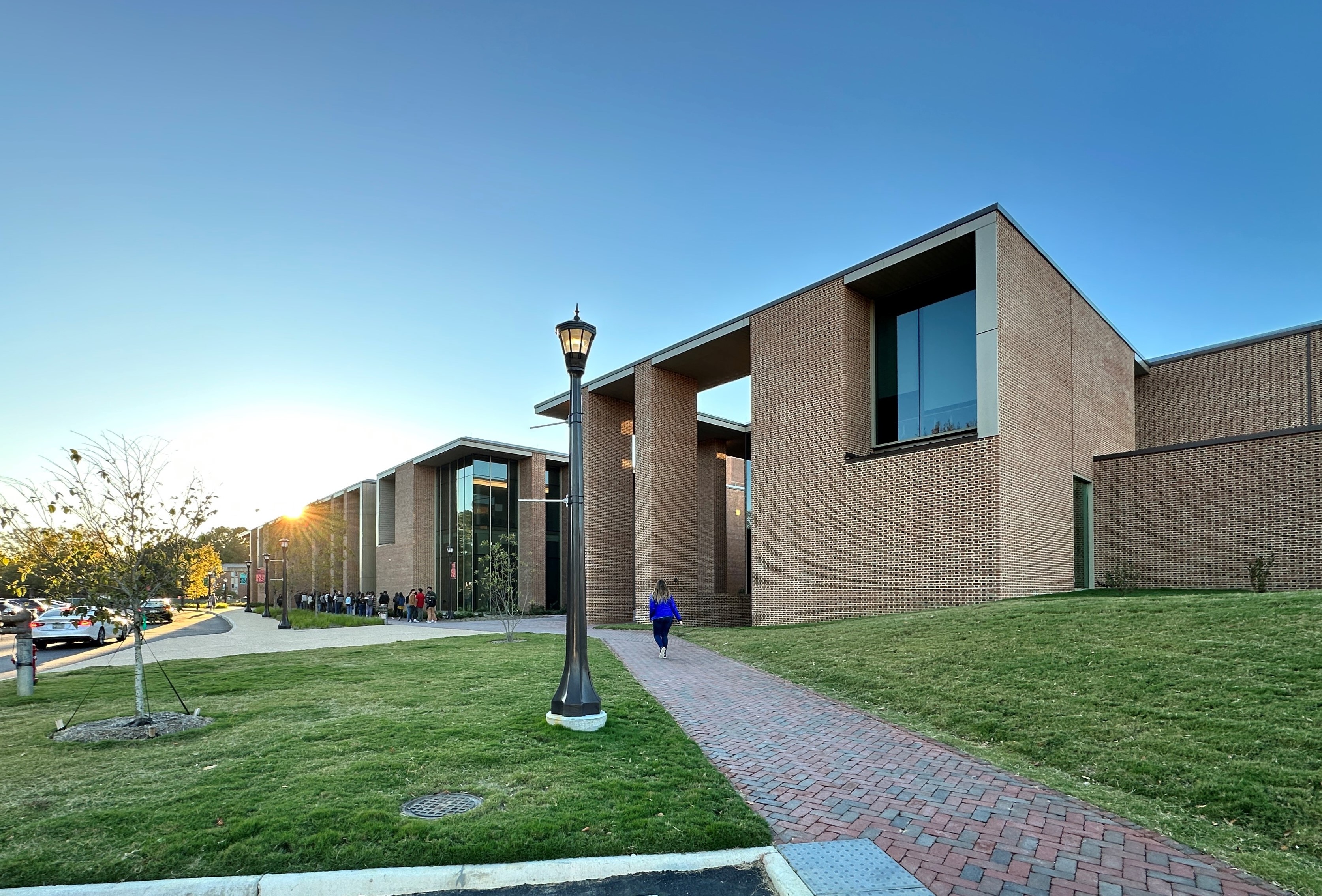 A woman in a blue shirt walks along a path in front of a brick and glass college performing arts center.