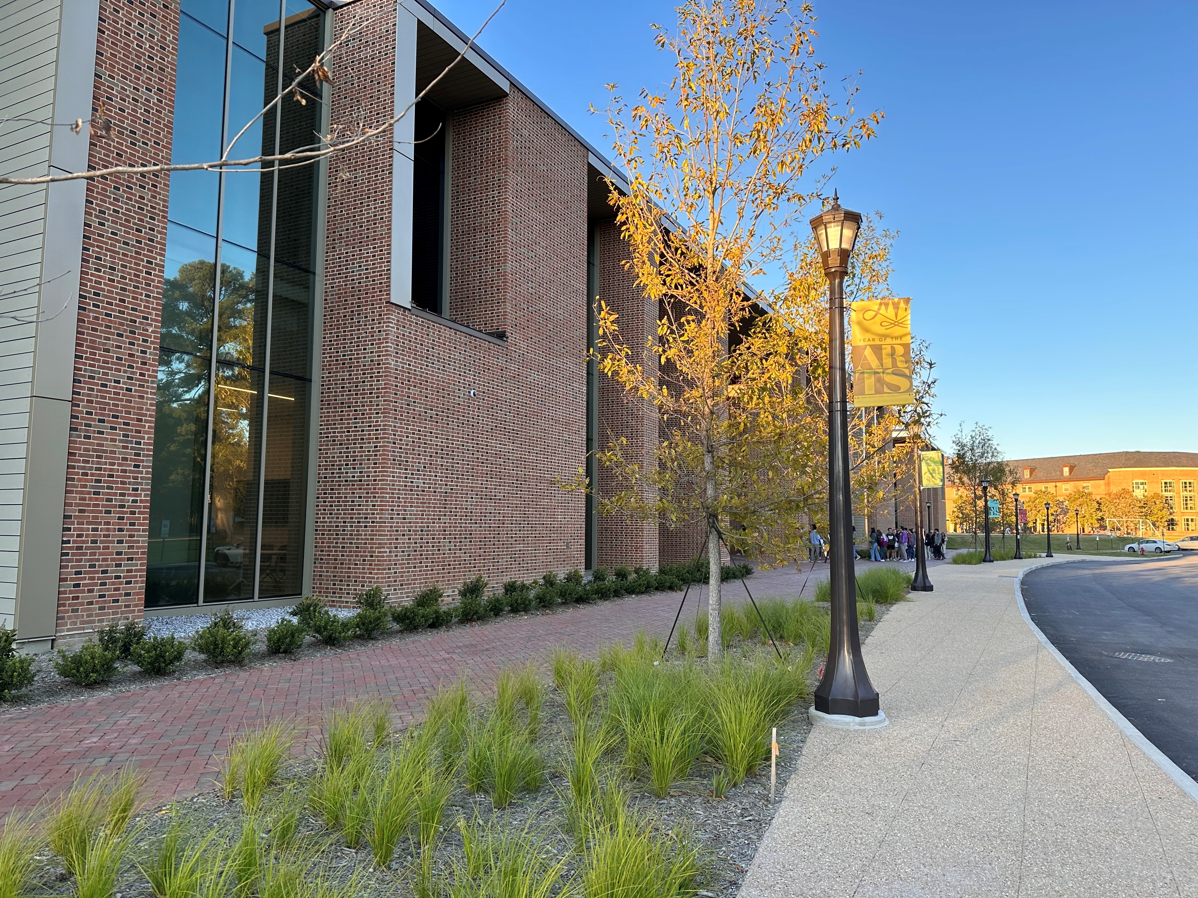 Light posts and newly planted trees and grasses line a walkway to a new brick and glass building.