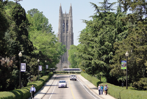 The road leading into Duke University, with people walking on sidewalks and the campus in the background.