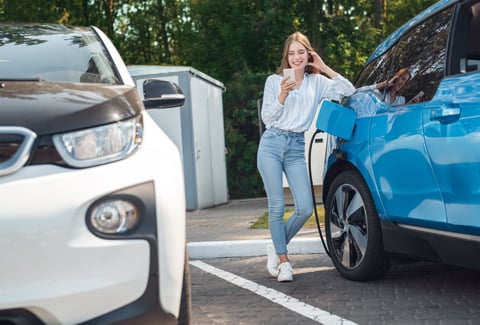 Young woman leaning on electric vehicle while it charges.