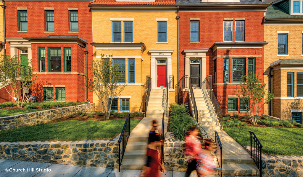 A group of people walking past stairs leading to a row of multi-story red and yellow brick townhomes.