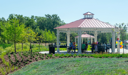 People gather inside a red-roofed picnic pavilion near landscaped plantings and vegetation.