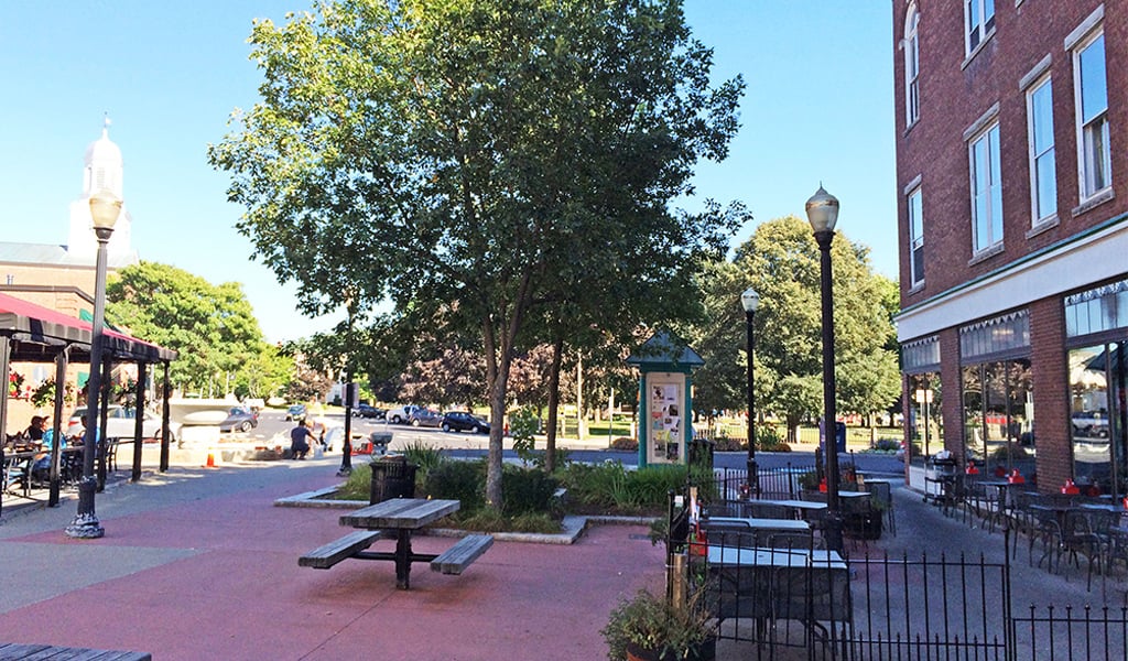Picnic tables and benches sit in the shade of a tree in a town square outside a brick building.