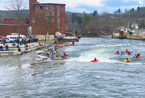 Numerous kayakers in multi-colored kayaks paddle in river rapids near a spectator viewing area.