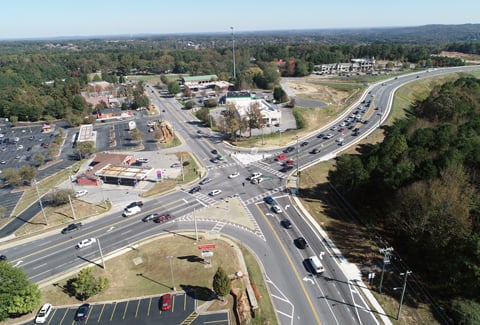An aerial photograph depicts a multi-lane roadway interchange with vehicles traveling on it and trees and buildings located beside the roadway.
