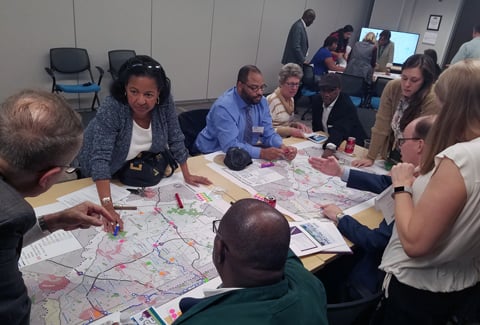 A diverse group of people sit and stand around a table looking at two maps of transit routes. 
