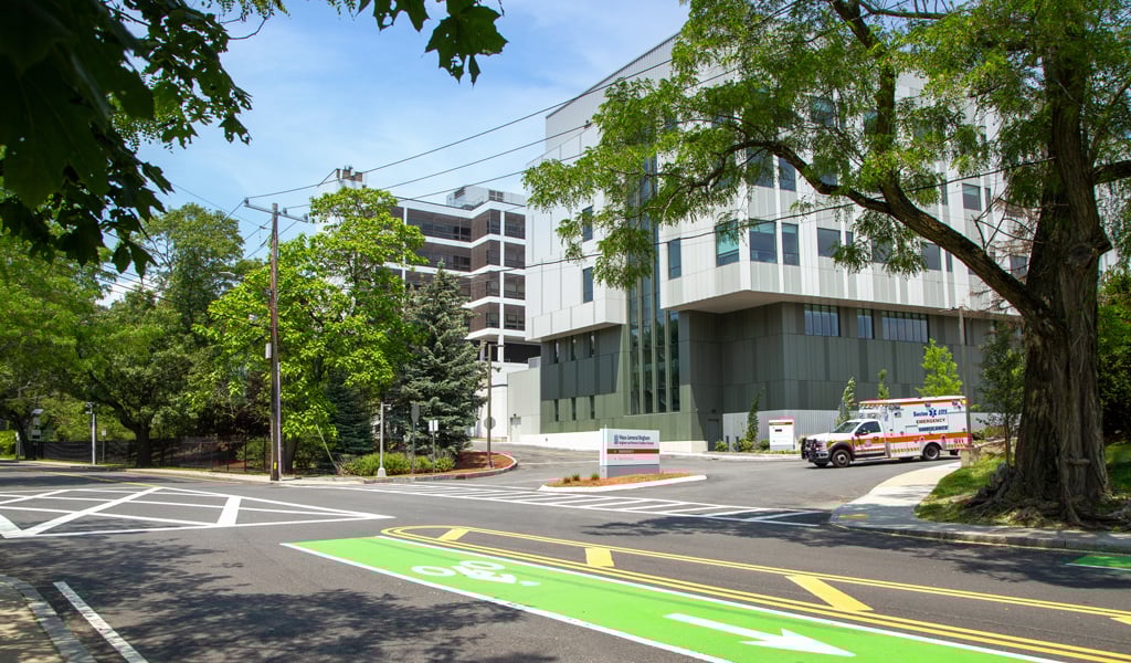 Modern hospital building with signage and ambulance, under a blue sky surrounded by a paved road with green bike lane and trees.