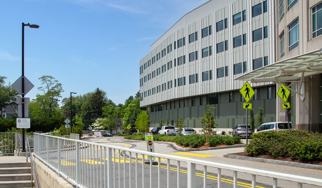 Side view of a modern hospital building with windows, pedestrian crossing and signage, sidewalk with railing, surrounded by greenery and parked cars along the road.