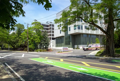 Modern hospital building with signage and ambulance, under a blue sky surrounded by a paved road with green bike lane and trees.