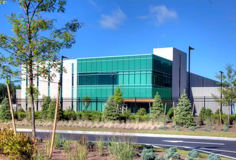 A modern office building with a green-tinged glass facade, surrounded by landscaped plants under a clear blue sky.