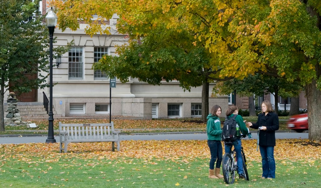Three people stand on a lawn surrounded by a large tree with autumn foliage and a brick academic building in the background.