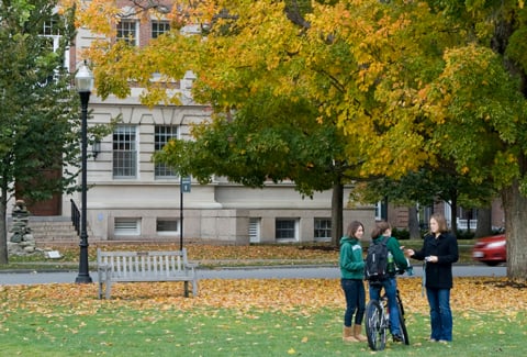 Three people stand on a lawn surrounded by a large tree with autumn foliage and a brick academic building in the background.