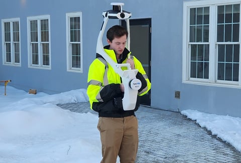A person wearing a high-visibility vest holding a surveying instrument (NavVis VLX) outside a building with snow on the ground.