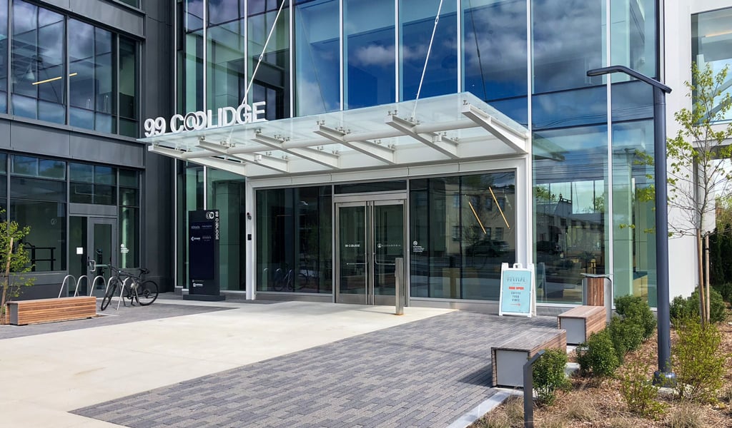 Modern glass industrial building front entrance labeled "99 Coolidge" with benches, bike rack, and plants.