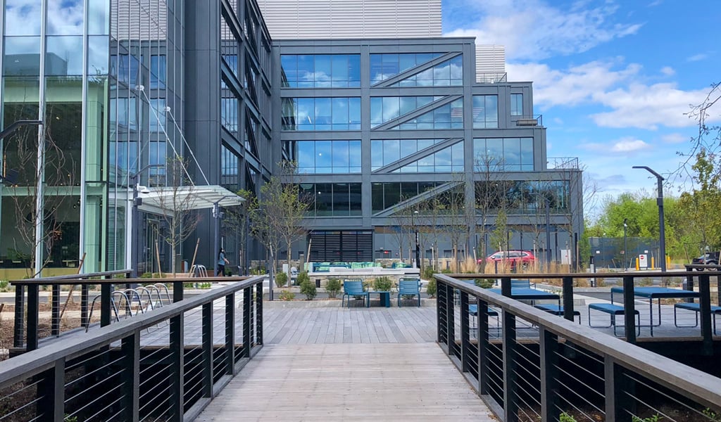 Modern glass industrial building with a wooden walkway and outdoor seating area under a cloudy blue sky