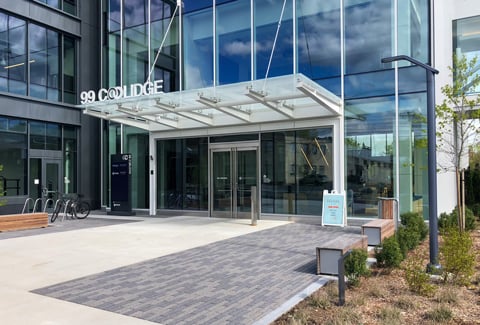 Modern glass industrial building front entrance labeled "99 Coolidge" with benches, bike rack, and plants.