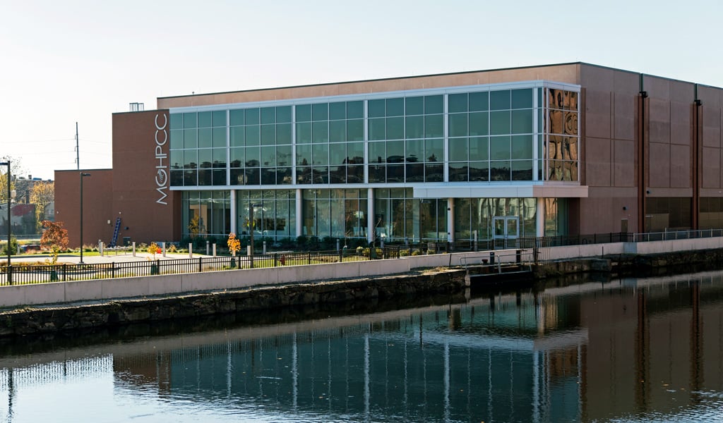 Modern glass-front building with fall trees, a road and tall vertical sign on a sunny day.