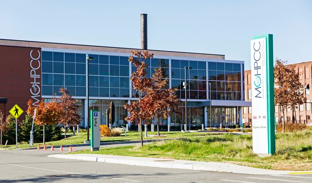 A modern glass-front building situated next to and reflected in a calm canal on a clear, sunny day.