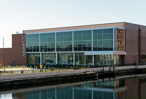 Modern glass-front building with fall trees, a road and tall vertical sign on a sunny day.