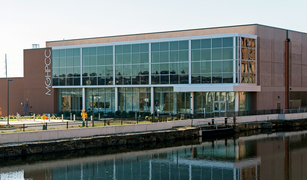 Modern glass-front building with fall trees, a road and tall vertical sign on a sunny day.