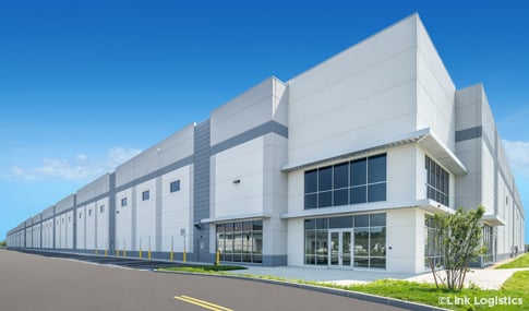 Front entry of a large light gray warehouse with glass windows and doors on a sunny day with blue skies.