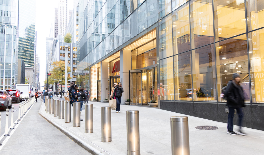 People walking on a city sidewalk beside a modern glass office building with bollards lining the street.