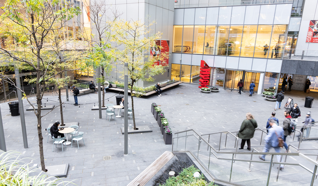 Modern courtyard with tables, benches, trees and people near a modern office building.
