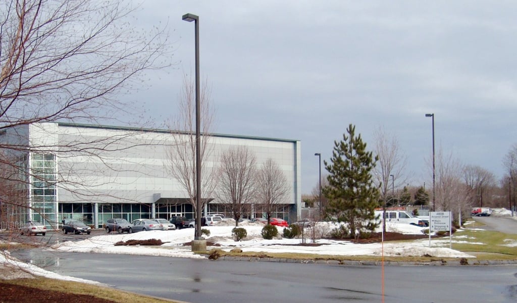 Modern industrial building with glass exterior, wet pavement, and bare trees under gray sky.