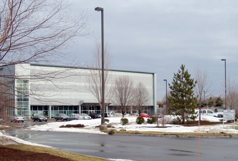 Modern industrial building with glass exterior, wet pavement, and bare trees under gray sky.