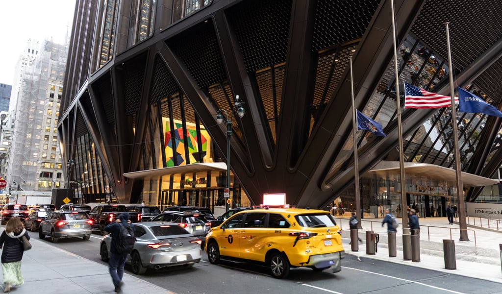 Yellow taxi and cars driving past the JPMorgan Chase building in Manhattan, with people on the sidewalk.