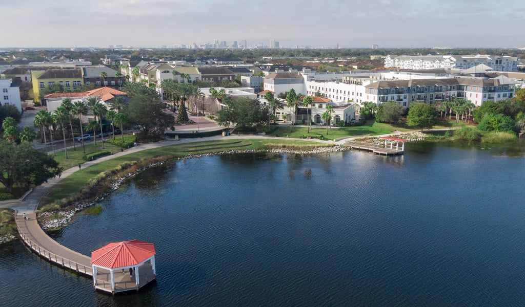 Aerial view of a lakeside boardwalk and gazebo beside a landscaped residential neighborhood with multi-story buildings.