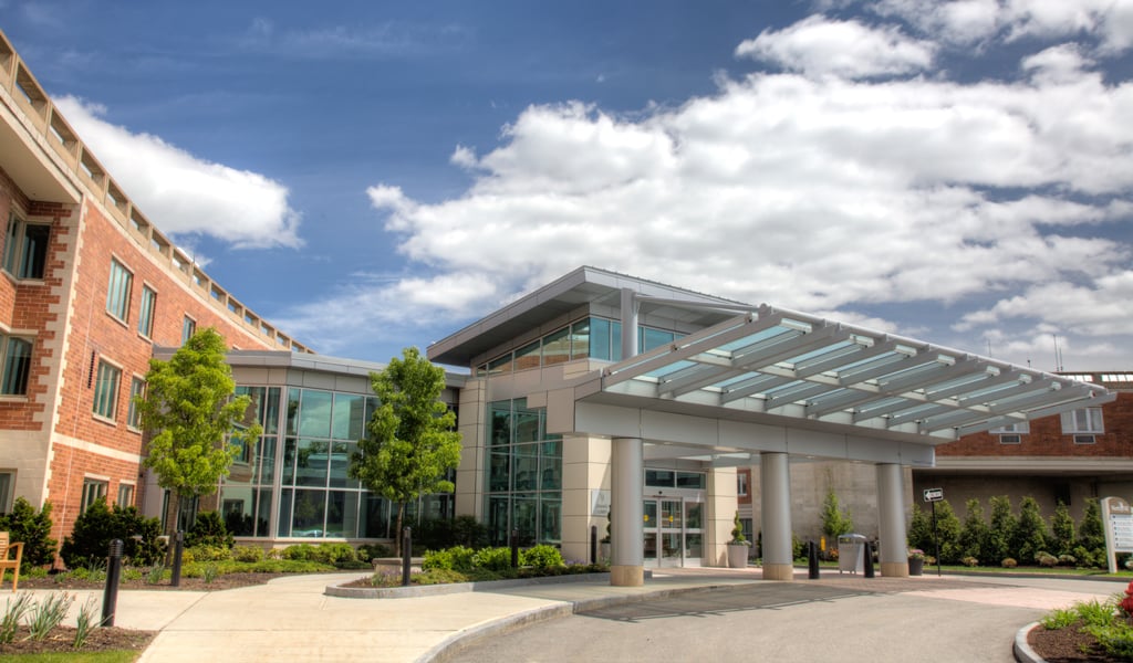 Modern building entrance with glass canopy, brick and glass facade, trees, under a cloudy sky.