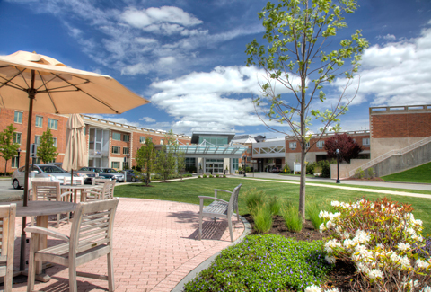 Sunny courtyard with patio tables, lawn, flowers, in the foreground of senior living buildings with a cloudy sky. 