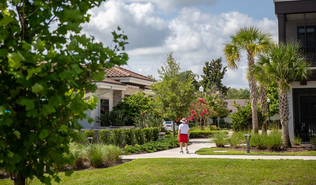 A resident at the Mayflower Retirement Community facility walking on a pathway adjacent to gardens and senior residences. 