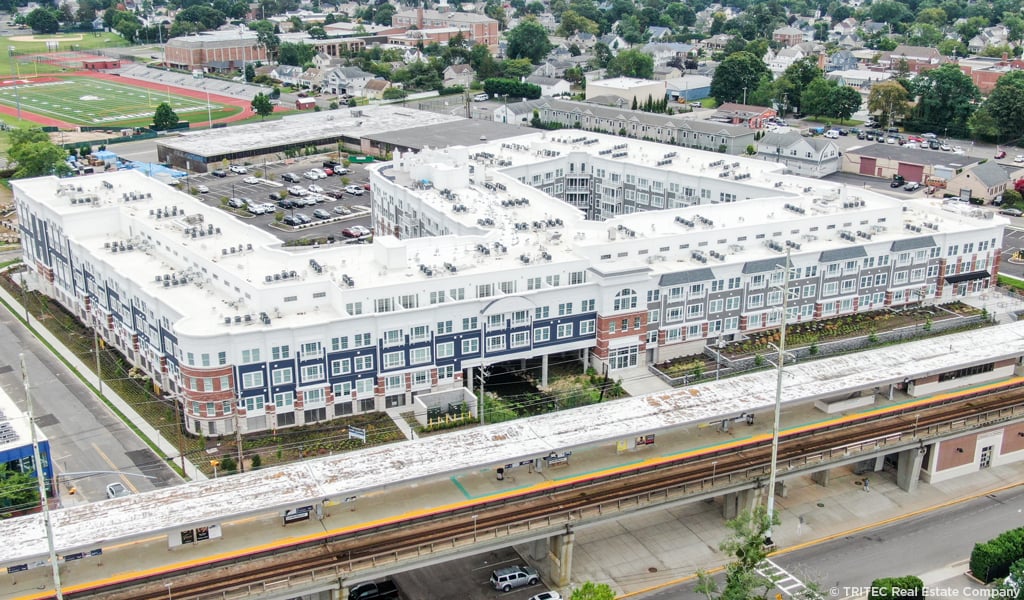 Aerial view of a large residential complex near a transit station with parking and sports fields in the background. 