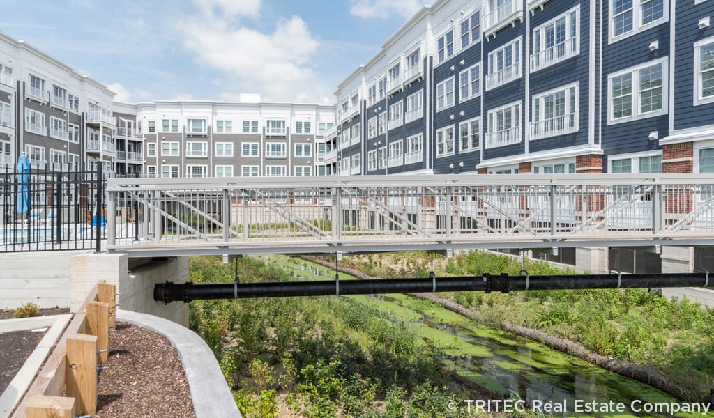 Restored stream flowing under a bridge beside modern apartments, highlighting urban daylighting of natural waterways.