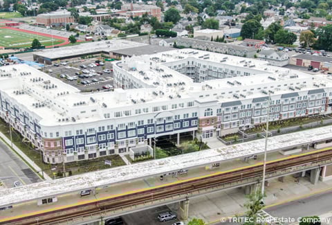 Aerial view of a large residential complex near a transit station with parking and sports fields in the background. 
